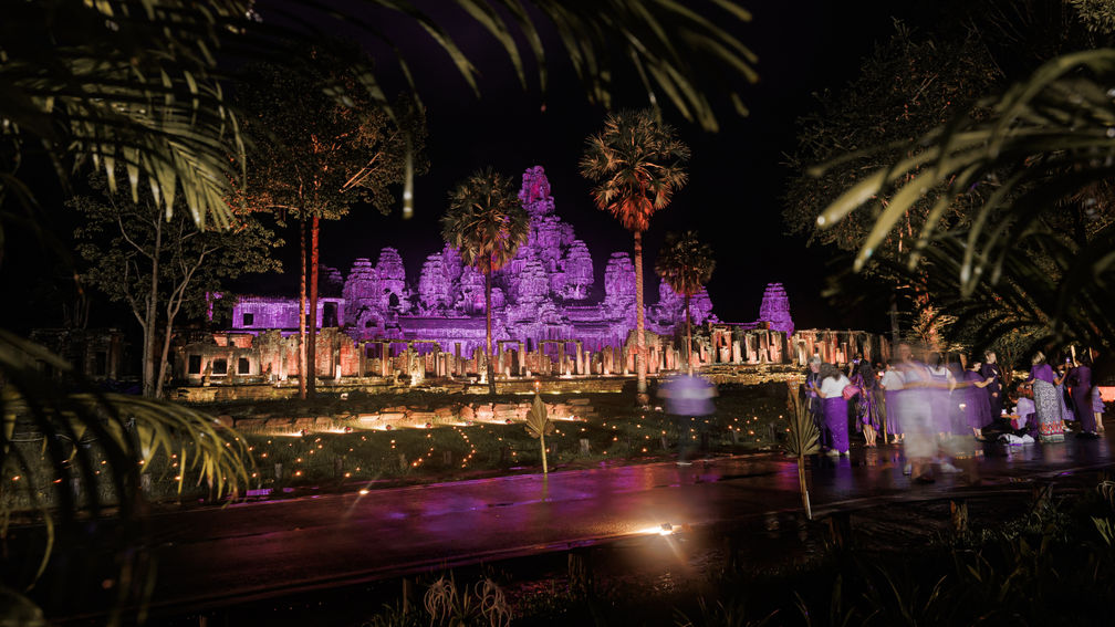Night scene of a purple-lit ancient temple ruins reflected in water.