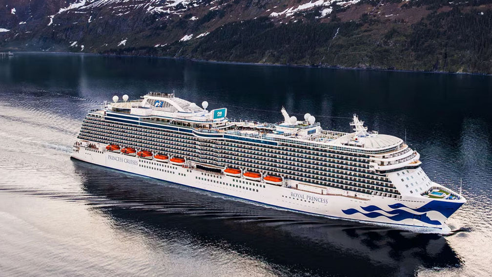 A large white cruise ship sails through calm water beside snow-capped cliffs.