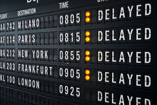 A large airport departure board shows delayed flights with yellow indicators.