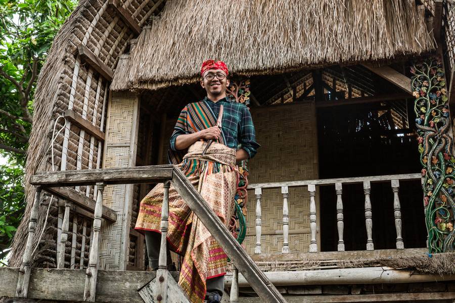Young man tourist posing with traditional Lombok costume in front of Traditional Sasak house