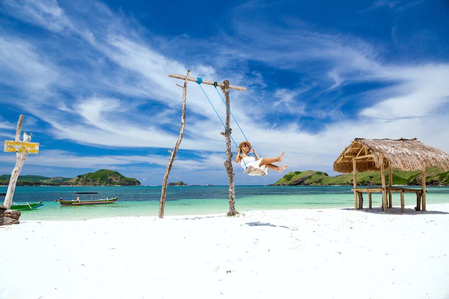 Woman in white dress and hat swinging at tropical beach, sunny day, good weather