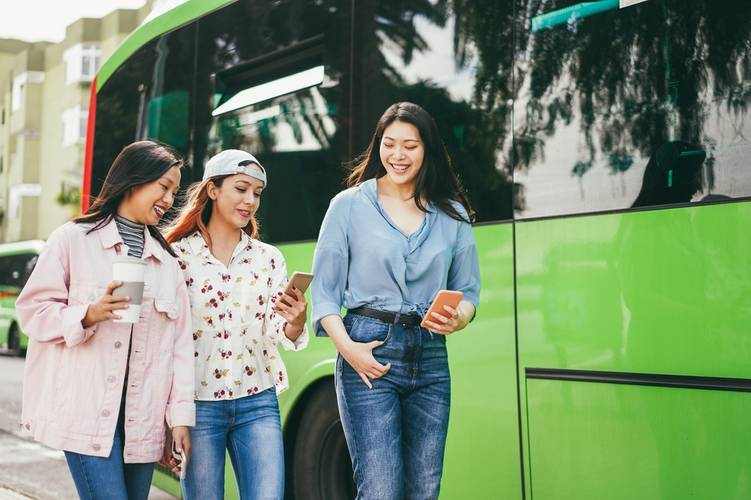 Happy young asian friends using mobile phones at bus station 