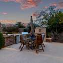 Cook and entertain in style with this stacked-stone outdoor kitchen featuring a stainless BBQ, beverage fridge, and counter seating. Set against a lush desert backdrop, this setup delivers function and flair in one refined space.