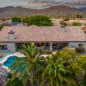 This aerial shot captures the full scale of the resort-style backyard, featuring a sparkling pool, covered patio, mature palms, and mountain views. A true desert retreat perfect for entertaining or quiet relaxation.