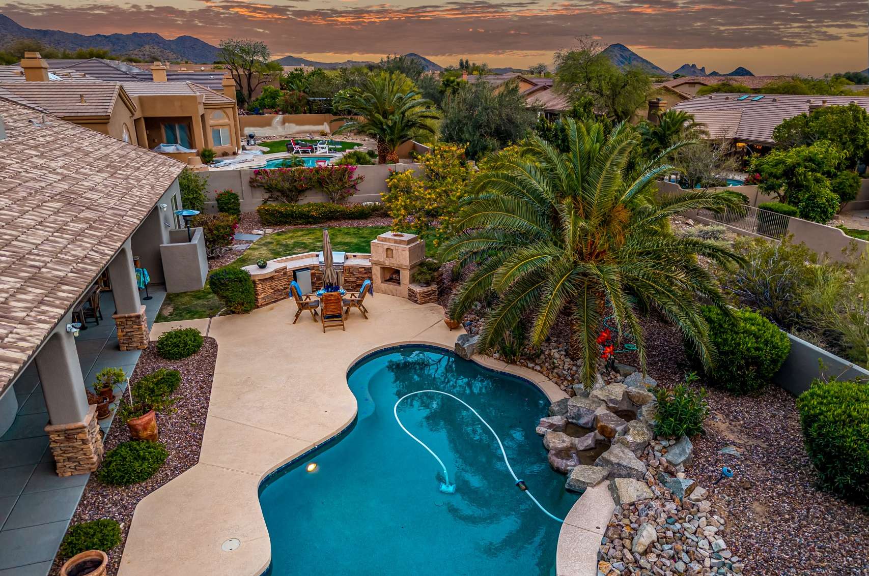27 of 65. Overlook a breathtaking backyard with a lagoon-style pool, built-in BBQ, and lush palm trees all set beneath colorful skies and scenic mountain silhouettes.