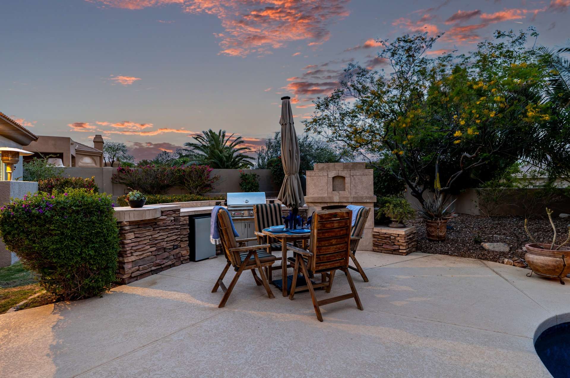 44 of 65. Cook and entertain in style with this stacked-stone outdoor kitchen featuring a stainless BBQ, beverage fridge, and counter seating. Set against a lush desert backdrop, this setup delivers function and flair in one refined space.