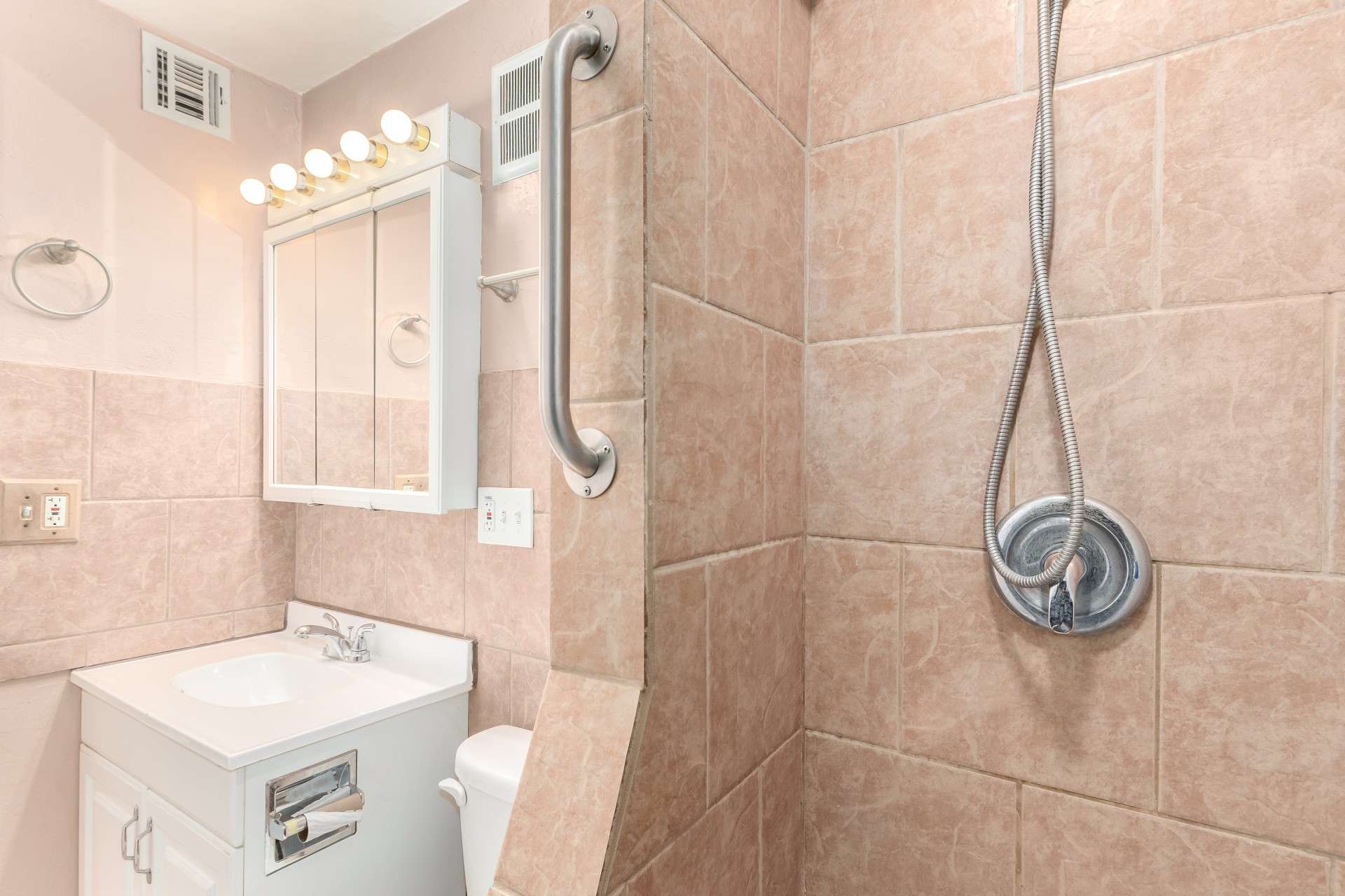 21 of 26. This well-designed bathroom features a fully tiled walk-in shower with a handheld showerhead, built-in grab bars, and modern lighting. The compact yet efficient vanity offers ample storage, making it both stylish and functional in this North Central Phoenix home.