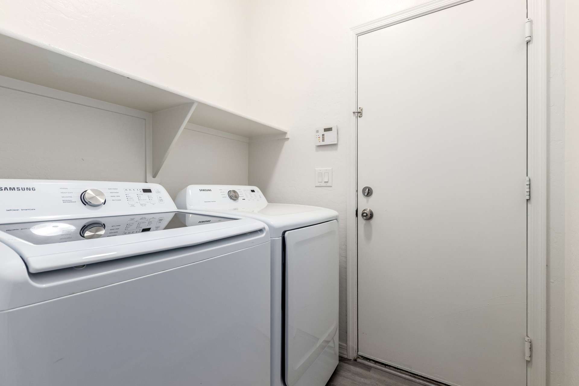 27 of 37. This functional laundry room includes Samsung top-load washer and dryer units, built-in shelving for efficient storage, and direct access to the garage. Clean finishes and neutral tones make laundry tasks effortless in this thoughtfully designed utility space.