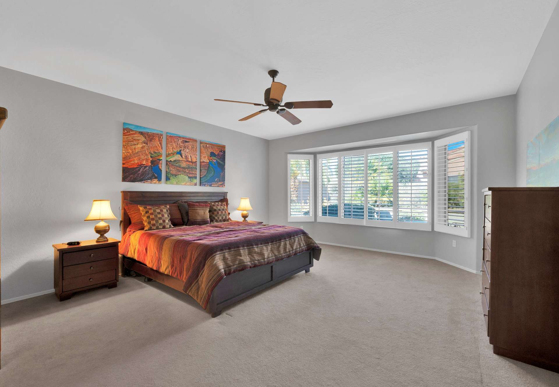 6 of 13. Unwind in this elegant primary bedroom featuring a king-sized bed, bay windows, ceiling fan, and soothing tones. A peaceful setting for a restful night during your Surprise stay.